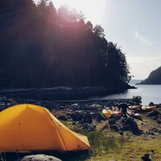 Waterside campsite with a yellow tent by a calm lake, surrounded by trees and mountains in natural sunlight.